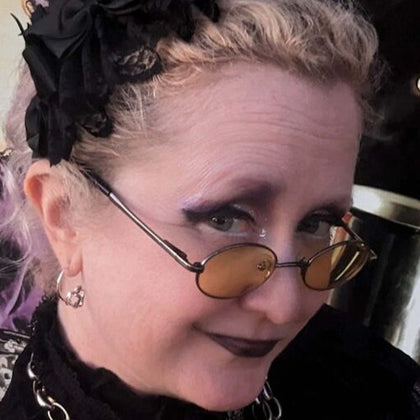 A person with styled blonde hair, wearing yellow-tinted glasses, black attire, and dark makeup, looking at the camera.