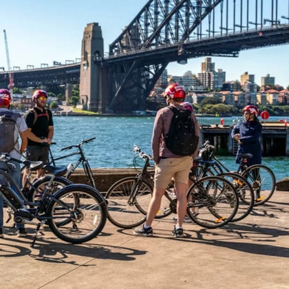 A group of cyclists with helmets gather near bicycles on a waterfront promenade with a large bridge and cityscape in the background.