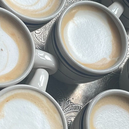 Several cups of cappuccino with frothy milk tops arranged closely together on a patterned metal tray.
