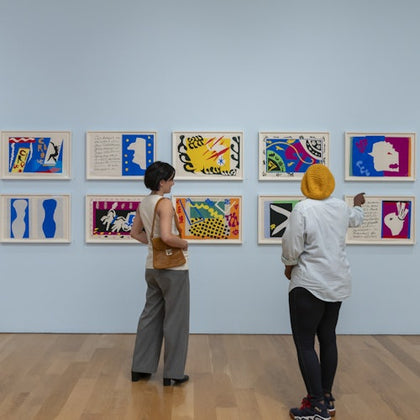 Two people stand in an art gallery, observing a wall of colorful framed artworks displayed on a light blue wall.