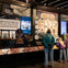 A museum exhibit displaying various items including a quilt, plates, menorah, and artifacts. Visitors observe the display.