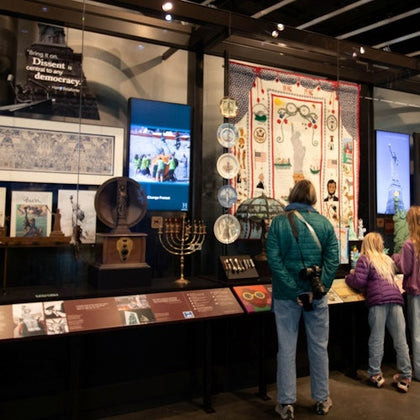 A museum exhibit displaying various items including a quilt, plates, menorah, and artifacts. Visitors observe the display.