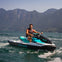 Man riding a teal and black Sea-Doo jet ski on a lake with mountains in the background on a clear day.