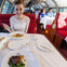 A woman in a white lace top dines on a red train with large windows, enjoying a meal and drinks. Other passengers are in the background.