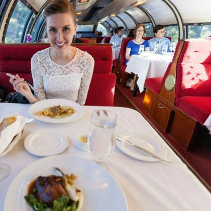 A woman in a white lace top dines on a red train with large windows, enjoying a meal and drinks. Other passengers are in the background.