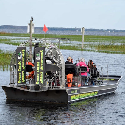 A group of people on an airboat with "Boggy Creek Airboat Adventures" signage, navigating through a marshy area.