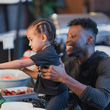 A smiling man holds a young child as they reach for food on a buffet table, with trays of fruit visible in the background.