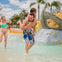 A man carries a child on his back in a water play area with slides and palm trees, while a woman stands nearby smiling.