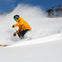 A skier in a yellow jacket and black pants skiing down a snowy slope, creating a spray of snow under a clear blue sky.