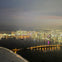 Aerial view of a city skyline at night with illuminated buildings, a bridge over water, and reflections of lights on the surface.