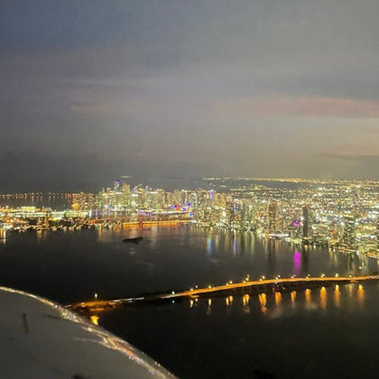 Aerial view of a city skyline at night with illuminated buildings, a bridge over water, and reflections of lights on the surface.