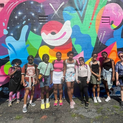 A group of people standing in front of a colorful graffiti mural on a sunny day.