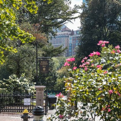 A park entrance with a sign reading "Public Garden 1837," surrounded by trees, flowers, and a distant view of buildings.
