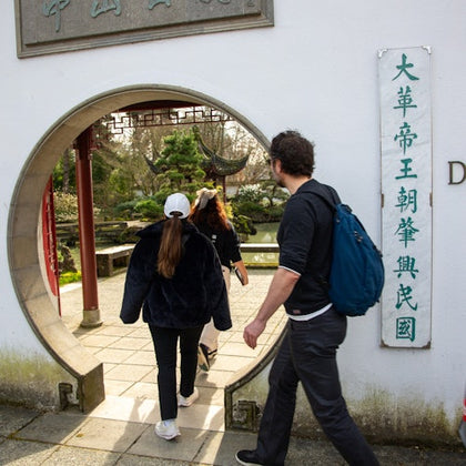 People walk through a circular entrance in a white wall with Chinese characters, leading to a garden with greenery and traditional architecture.