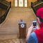 A person stands at a podium in front of a grand staircase, while another person in a red hat takes a photo with a smartphone.