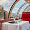 A table set for two with glassware and utensils in a scenic restaurant with large curved glass windows and red seating.