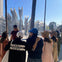 A group of people stands near tall lamp posts in an outdoor area on a sunny day, with palm trees and a Metro sign visible.