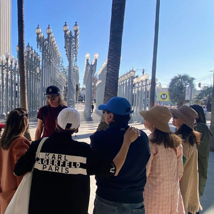 A group of people stands near tall lamp posts in an outdoor area on a sunny day, with palm trees and a Metro sign visible.