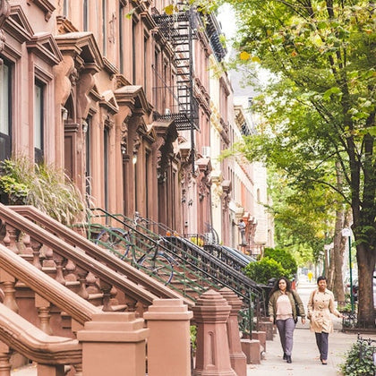 Two people walking on a sidewalk lined with brownstone buildings and trees on a sunny day.