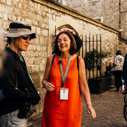 A woman in an orange dress is smiling and talking to a person wearing a hat and sunglasses on a cobblestone street by a stone wall.