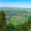 Scenic view from a hilltop overlooking a town surrounded by green fields, trees, and distant hills under a clear blue sky.