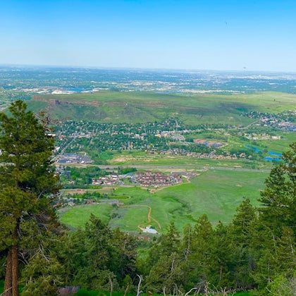 Scenic view from a hilltop overlooking a town surrounded by green fields, trees, and distant hills under a clear blue sky.