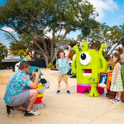 A man photographing a child posing next to a green cartoonish character sculpture outdoors on a sunny day.