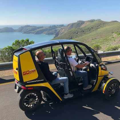 Two people riding a small yellow electric vehicle labeled "GoCar Tours" on a coastal road with scenic ocean views.