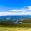 A scenic view of a forested valley with a small lake, surrounded by distant mountains under a clear blue sky.