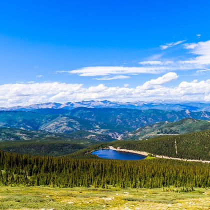 A scenic view of a forested valley with a small lake, surrounded by distant mountains under a clear blue sky.