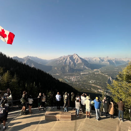 People stand on a viewing platform overlooking a vast mountainous landscape with a Canadian flag flying.