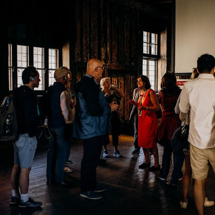 A group of people attentively listening to a presenter in a dimly lit room with wooden floors and large windows.