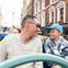 A man and a boy smiling at each other while riding a double-decker bus in a city with brick buildings in the background.