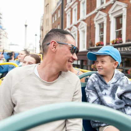 A man and a boy smiling at each other while riding a double-decker bus in a city with brick buildings in the background.