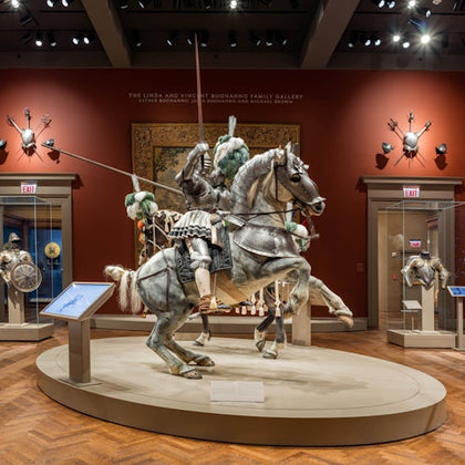 A museum exhibit displaying a horse and rider in full medieval armor on a pedestal, surrounded by arms and armor displays.