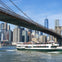 A sightseeing boat passes under the Brooklyn Bridge with the New York City skyline in the background.