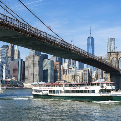 A sightseeing boat passes under the Brooklyn Bridge with the New York City skyline in the background.