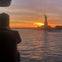 A person views the Statue of Liberty at sunset from a waterfront, with the statue silhouetted against a colorful sky.