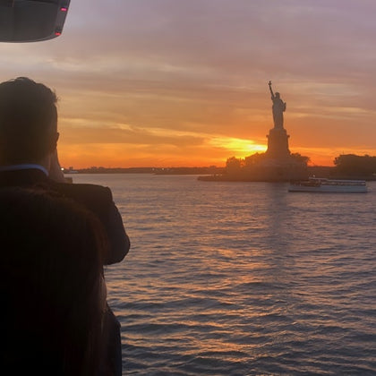 A person views the Statue of Liberty at sunset from a waterfront, with the statue silhouetted against a colorful sky.