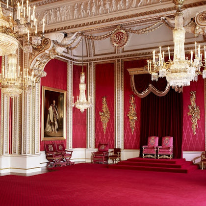 A lavish red and gold room with chandeliers, ornate decorations, a painting, and two armchairs on a small stage.