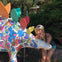 A woman smiling next to a colorful, decorated stegosaurus statue with cartoonish designs and vibrant plates. Trees and bushes in the background.