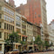 A city street lined with historic multi-story buildings, trees, parked cars, and pedestrians, under an overcast sky.