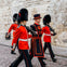 Three guards in red uniforms and bearskin hats march alongside a man in ceremonial dress by a stone wall.