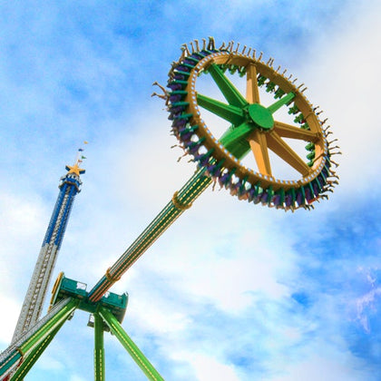 People riding a colorful carousel swing ride in an amusement park, with trees and a building in the background under a clear sky.