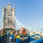 Tourists seated on the upper deck of an open-top bus crossing Tower Bridge under a clear blue sky.