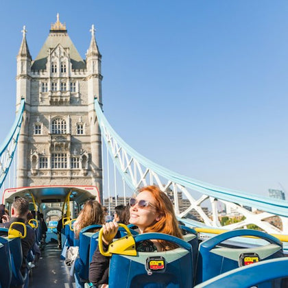 Tourists seated on the upper deck of an open-top bus crossing Tower Bridge under a clear blue sky.