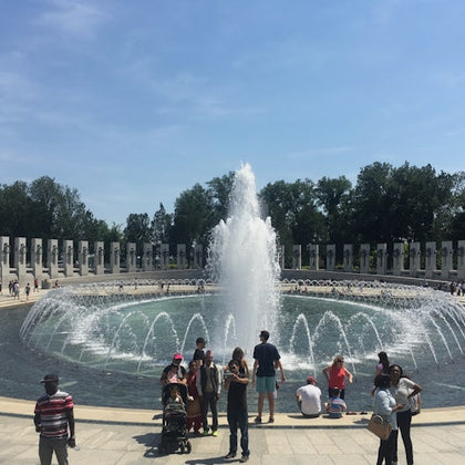 People gather around a large fountain with water jets in a circular pool, surrounded by a colonnade and greenery.