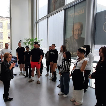 A group of people standing indoors near large windows, listening to a woman speaking. A poster is visible in the background.