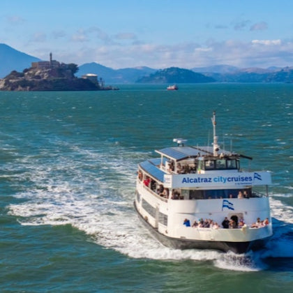 A ferry labeled "Alcatraz City Cruises" sails on water, with Alcatraz Island and a distant shoreline in the background.