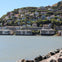 A hillside with numerous houses overlooks a calm body of water, with rocks lining the foreground under a blue sky.